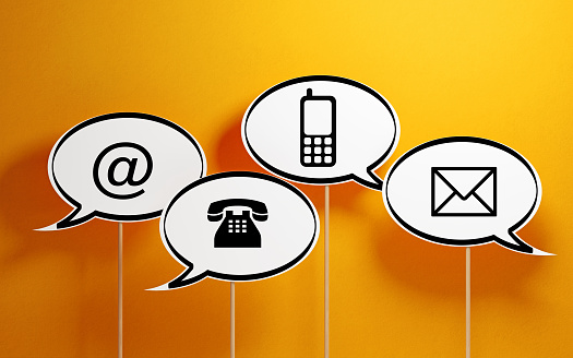 White chat bubbles with wooden sticks on yellow background. There are various contact us symbols on the chat bubbles. Horizontal composition with copy space.